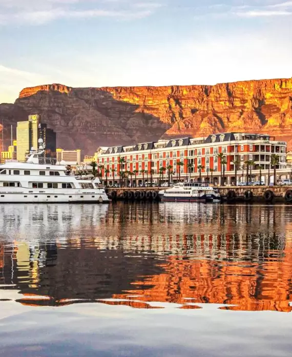 Cape Grace Hotel at the V&A Waterfront in Cape Town, South Africa, with the iconic Table Mountain illuminated at sunset in the background.