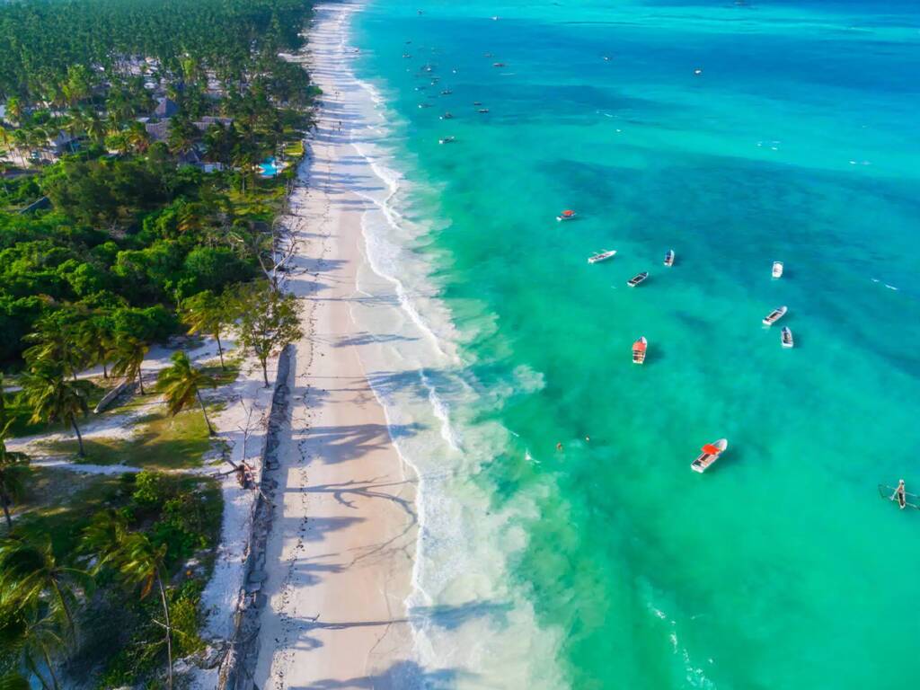 Zaznibar’s Paje Beach with long stretches of white sandy shoreline with palm trees and boats.