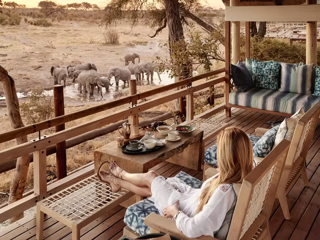 A traveler, enjoying breakfast on her private deck, watches an elephant herd drink water on a luxury African safari trip in Botswana. 