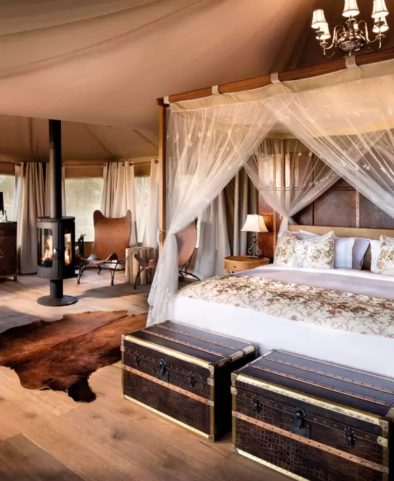 Elegant tented suite at One Nature Nyaruswiga in Tanzania, featuring luxurious decor with a canopy bed, vintage trunks, and a cozy fireplace.