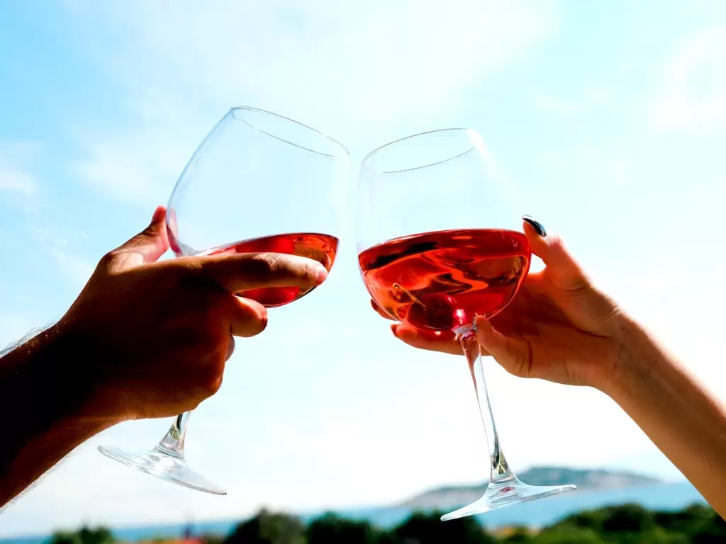 A couple’s hand is holding glasses of wine, doing cheers, and raising a toast to their love on a honeymoon trip.