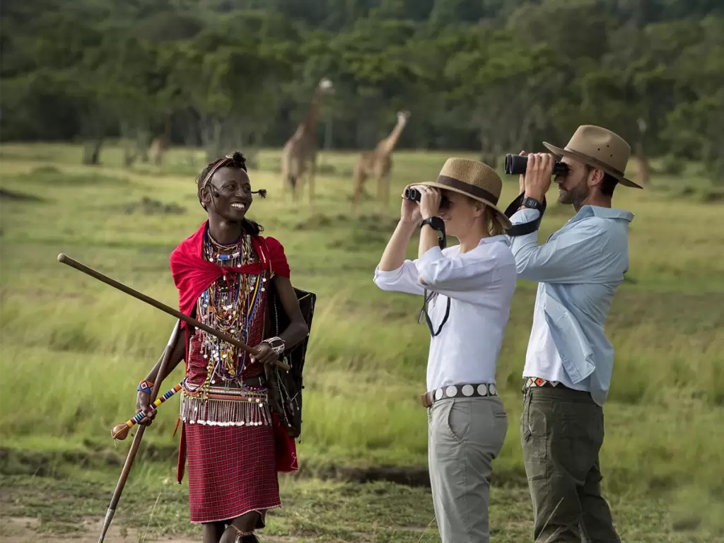 A couple on a luxury African walking safari, led by a Masai guide in the African savannah with giraffes in the backdrop.
