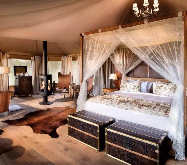 Elegant tented suite at One Nature Nyaruswiga in Tanzania, featuring luxurious decor with a canopy bed, vintage trunks, and a cozy fireplace.